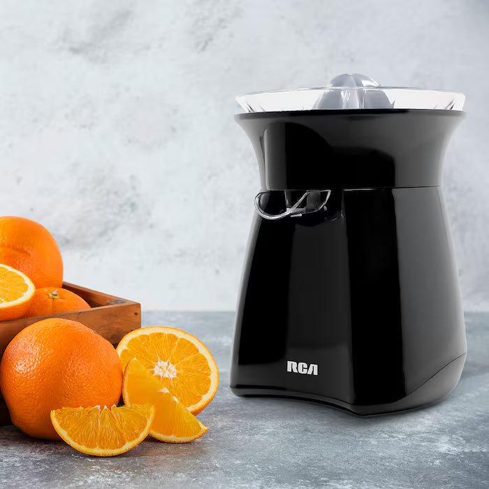 Exprimidor de cítricos negro RCA con diseño compacto y boquilla anti goteo junto a naranjas frescas cortadas.
