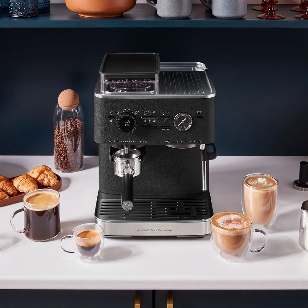 Cafetera semiautomática KitchenAid negra con control de presión, molinillo integrado y espumador de leche, en barra blanca.